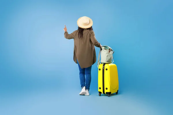 Achteraanzicht van jonge zwarte vrouw met bagage wijzend, het kiezen van toeristische tour op blauwe studio achtergrond, kopieerruimte — Stockfoto