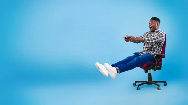 Возбужденный молодой черный парень с джойстиком сидит на стуле, играет в видеоигру на синем фоне, баннер с копировальным пространством — стоковое фото