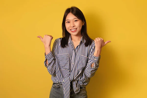 Joyful jovem mulher asiática em casual desgaste mostrando polegares para cima com as duas mãos, sorrindo para a câmera no fundo amarelo — Fotografia de Stock