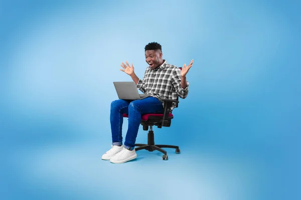 신나는 젊은 흑인 남자 노트북 과 의자에 앉아, 블 루 스튜디오 배경에 성공이나 성취를 축하 — 스톡 사진
