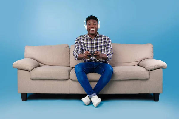 조이스틱 과 비디오 게임을 하고 헤드셋을 쓰고 소파에 앉아 푸른 스튜디오 배경 위에 앉아 있는 행복 한 젊은 흑인 남자 — 스톡 사진