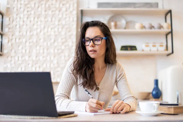 Freelance, gerente remoto, contabilidade doméstica. Mulher de óculos fazendo anotações na mesa da cozinha com laptop — Fotografia de Stock