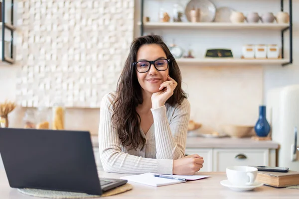Glad kvinna i glasögon och casual wear tittar och ler mot kameran, sitter med laptop vid bordet i köket interiör — Stockfoto
