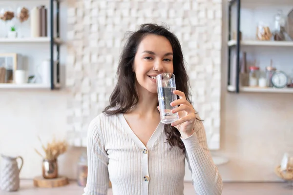 Fröhliche Frau trinkt sauberes Mineralwasser aus Glas und steht zu Hause in der Küche — Stockfoto