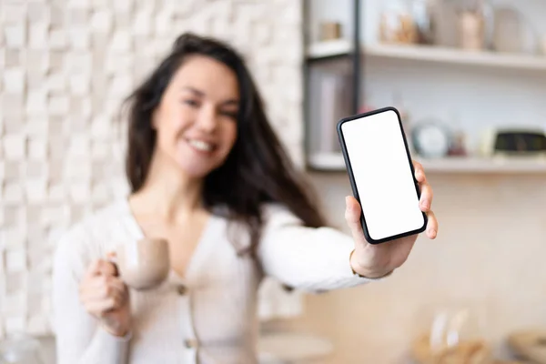 Mujer feliz de pie en el interior de la cocina, beber té y mostrar el teléfono celular moderno con pantalla en blanco, maqueta — Foto de Stock