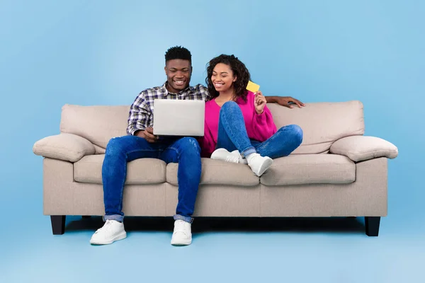 Pasangan kulit hitam belanja online melalui laptop dan kartu kredit, memesan sesuatu di internet, duduk di sofa, latar belakang biru — Stok Foto