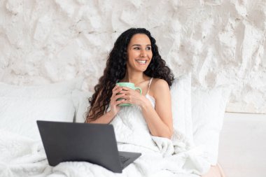 Neşeli, çekici, Avrupalı, uzun saçlı esmer kadın fincandan kahve içer, dizüstü bilgisayarla yatakta oturur.