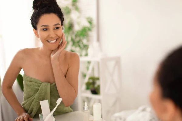 Hermosa joven en toalla mirándose en el espejo, tocándose la cara y sonriendo en casa, espacio libre — Foto de Stock