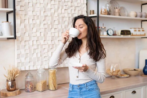 Glada kvinna i casual kläder dricka sin favorit varm dryck i vit kopp, stående i modernt kök interiör — Stockfoto