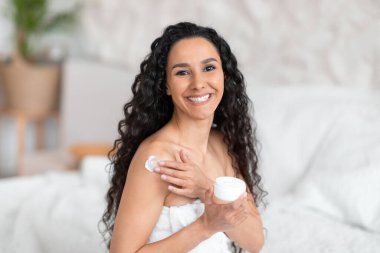 Gülümseyen, Avrupalı, uzun saçlı, uzun saçlı esmer kadın yatak odasında vücuduna krem sürüyor.