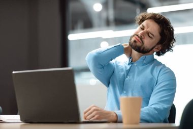 Masada oturan yorgun adam bilgisayarla boynuna masaj yapıyor.