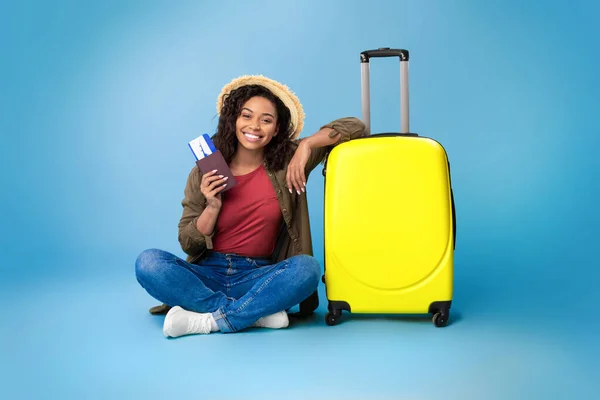 Preciosa joven negra con pasaporte y pasajes de avión, sentada cerca de la maleta de color amarillo brillante sobre fondo azul — Foto de Stock