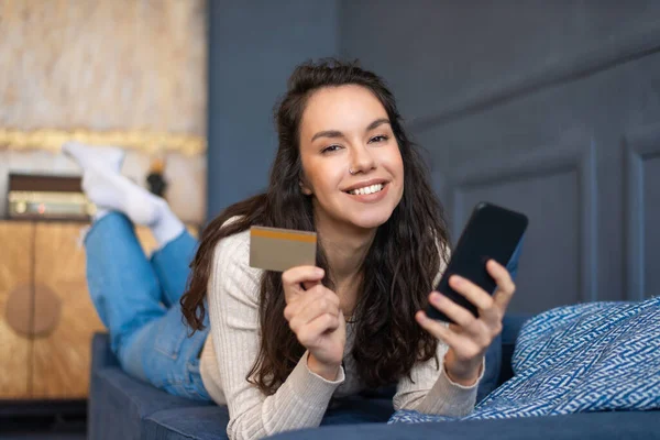 Concepto de pago online. Mujer alegre sosteniendo tarjeta de crédito y teléfono inteligente, acostado en el sofá y sonriendo a la cámara — Foto de Stock