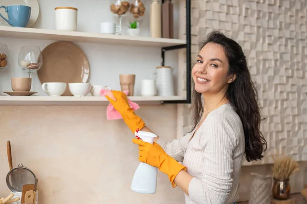 Ritratto di donna che indossa guanti di gomma protettiva che pulisce scaffali in cucina, tiene spray e usa un panno in microfibra — Foto Stock