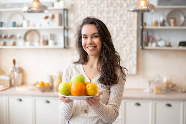 Happy woman holding plate with fresh fruits, looking and smile at camera, standing in kitchen interior — Stock fotografie