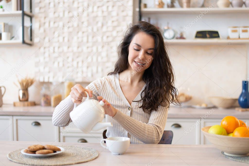 Cheerful woman in casual pouring tea in cup, sitting at table in kitchen interior, enjoying morning