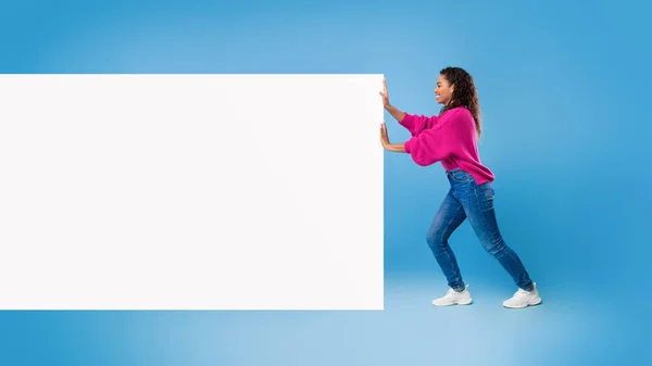Volle Länge einer jungen afroamerikanischen Dame, die ein leeres weißes Papierbanner mit Platz für Werbung auf blauem Hintergrund schiebt. Attrappe — Stockfoto