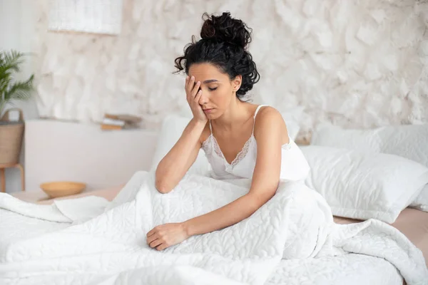 Κουρασμένος υπνηλία αρκετά χιλιετή ευρωπαϊκή μελαχρινή γυναίκα ξυπνάει μόνη της στη δουλειά, κάθεται στο κρεβάτι σε λευκό υπνοδωμάτιο — Φωτογραφία Αρχείου