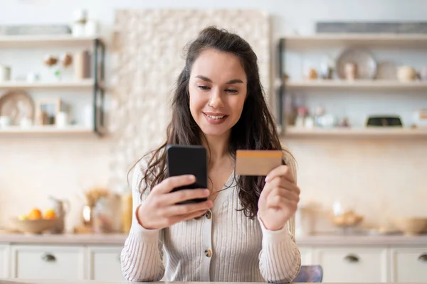 Aplicación de compras de comestibles. Mujer alegre usando tarjeta de crédito y teléfono celular, la compra de alimentos en línea mientras está sentado en la cocina — Foto de Stock