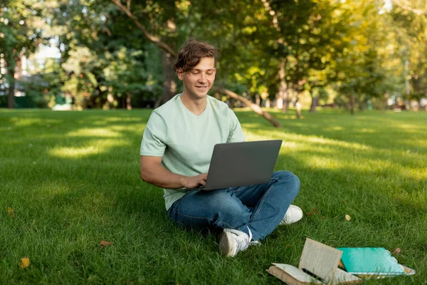 Веселый кавказский студент-мужчина с ноутбуком и учится онлайн, делает домашнюю работу на открытом воздухе, сидит на газоне — стоковое фото