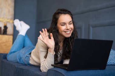Heyecanlı kadın videosu laptopla arıyor, bilgisayar kameralarına el sallıyor, oturma odasındaki kanepede uzanıyor.