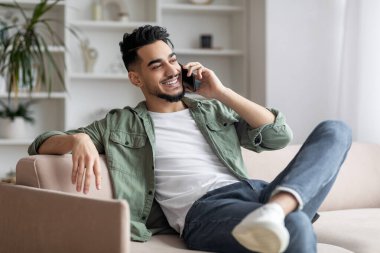 Neşeli Arap adam evdeki kanepede dinlenirken telefonla konuşuyor.