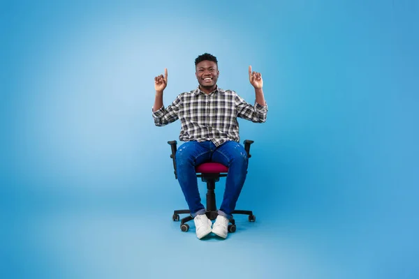Bonito jovem negro apontando dois dedos para cima no espaço vazio, sentado na cadeira no fundo do estúdio azul, mockup — Fotografia de Stock
