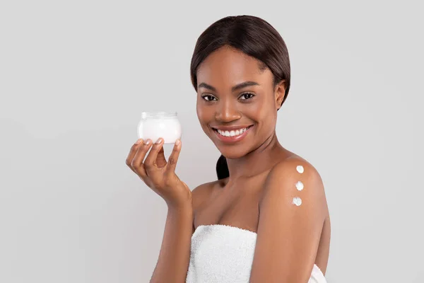 Smiling pretty millennial african american woman with perfect skin in towel with cream on shoulder showing jar — Stock Photo, Image