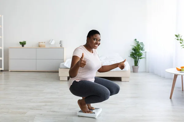 Feliz mujer afroamericana regordeta sentada en escamas, mostrando un gesto de pulgar hacia arriba, alcanzando su meta adelgazante en casa —  Fotos de Stock