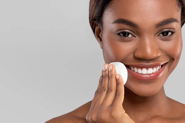 Sorrindo muito milenar afro-americano feminino limpando seu rosto com algodão pad, isolado em fundo cinza, close up — Fotografia de Stock