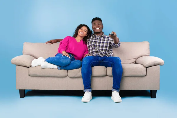 Opgewonden jong zwart paar met afstandsbediening zitten op de bank en tv kijken tegen een blauwe studio achtergrond — Stockfoto