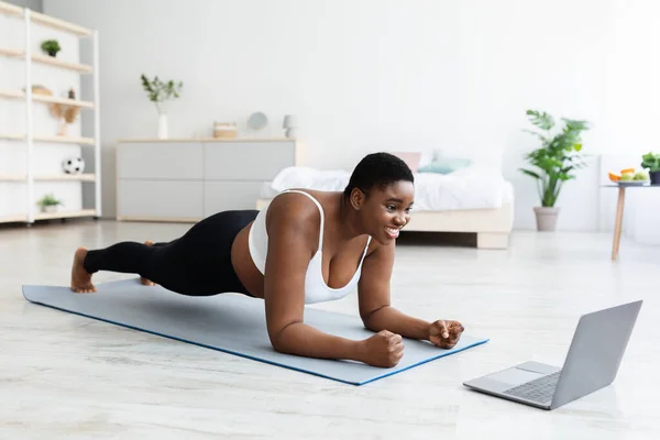 Online sports and weight loss. Overweight young black woman doing exercises in front of laptop at home, full length