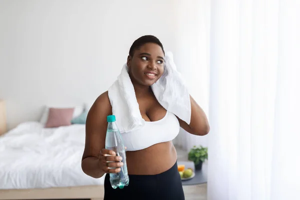 Ditambah ukuran wanita muda Afrika-Amerika dengan botol air membersihkan keringat dengan handuk setelah latihan di rumah — Stok Foto
