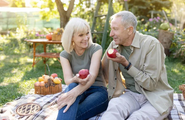 Hälsosam och lycklig pension. Positiva äldre makar som sitter på gräsmattan och äter äpplen, vilar i trädgården — Stockfoto