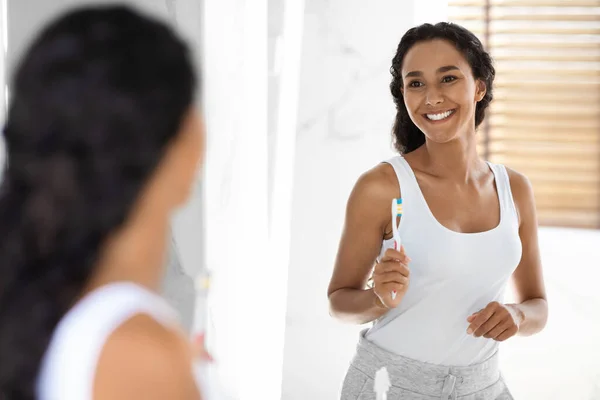 Приваблива тисячолітня жінка з зубною щіткою в руках стоїть біля дзеркала в ванній — стокове фото