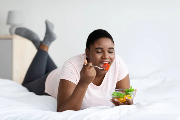 Wanita hitam gemuk makan salad sayuran lezat, berbaring di tempat tidur di rumah. gizi sehat untuk menurunkan berat badan — Stok Foto