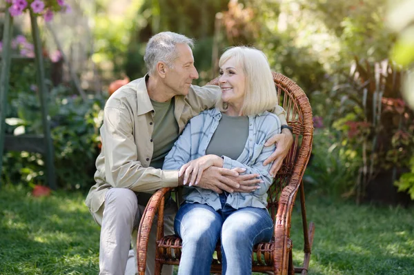 Šťastný starší pár těší čas na venkově, žena sedí v proutěném křesle a muž objímající ženu — Stock fotografie