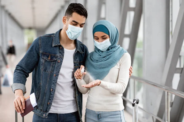 Viajes seguros. Joven pareja musulmana usando máscara médica aplicando desinfectante en el aeropuerto —  Fotos de Stock