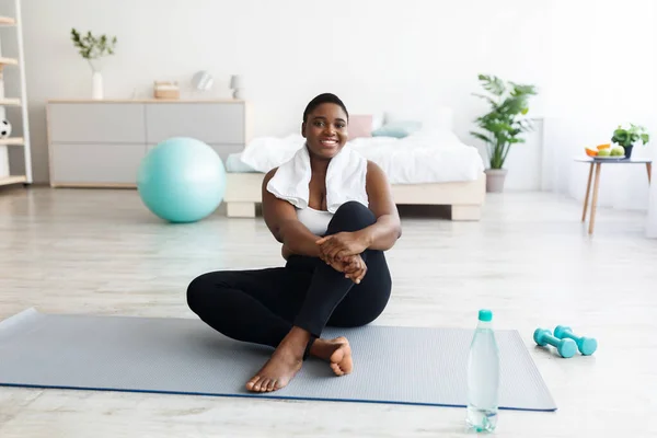 Sonriendo mujer negra joven con sobrepeso sentada en la esterilla de yoga, descansando después de la aptitud en el hogar, copiar espacio —  Fotos de Stock