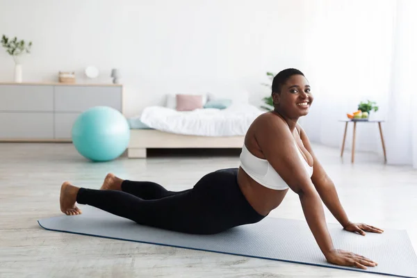 Толстая молодая черная леди растягивается на спортивном коврике, практикует йогу, пытается похудеть дома, полная длина — стоковое фото