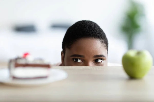 Plus size black woman choosing between cake and apple, peeking at food from under table — Stock Photo, Image