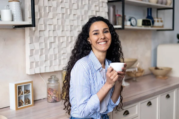 Portret van een vrolijke latijnse vrouw die thee drinkt en lacht naar de camera, in de keuken staat met een kopje in de handen, kopieerruimte — Stockfoto