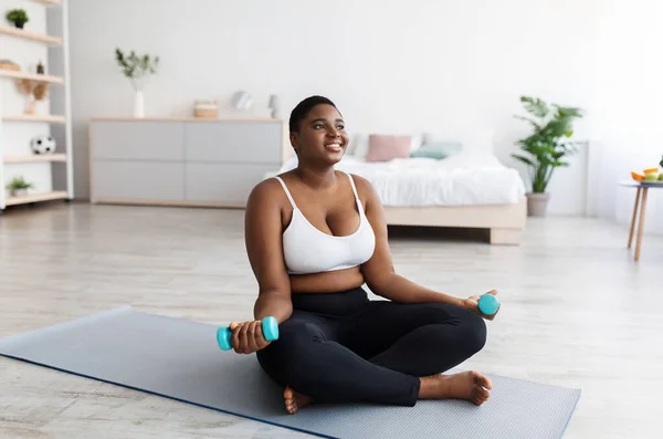 Mujer negra con sobrepeso en ropa deportiva sentada en una esterilla de yoga y haciendo ejercicio con pesas en casa, espacio para copiar — Foto de Stock