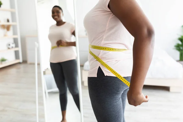 Обрізаний вид надмірної ваги молода чорна жінка вимірює талію стрічкою біля дзеркала вдома, вільний простір — стокове фото