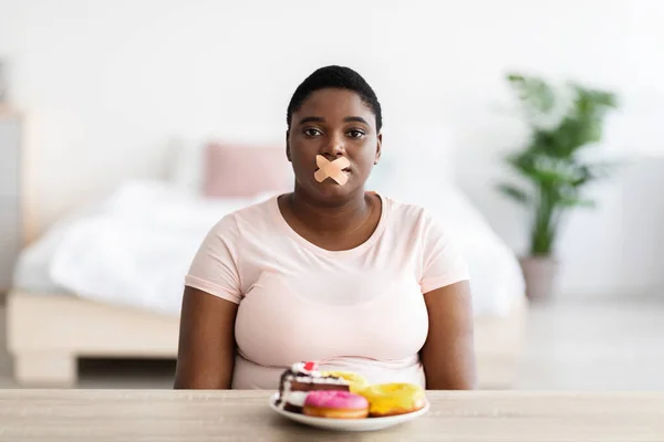 Upset curvy black woman with bandage on mouth sitting at table with plate of sweets, refusing desserts, losing weight