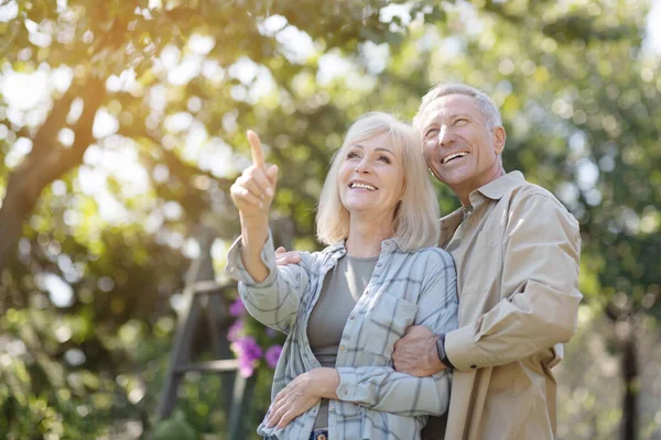 Tender senior spouses walking in their garden, enjoying warm spring day outdoors, embracing and looking away — Stock Photo, Image