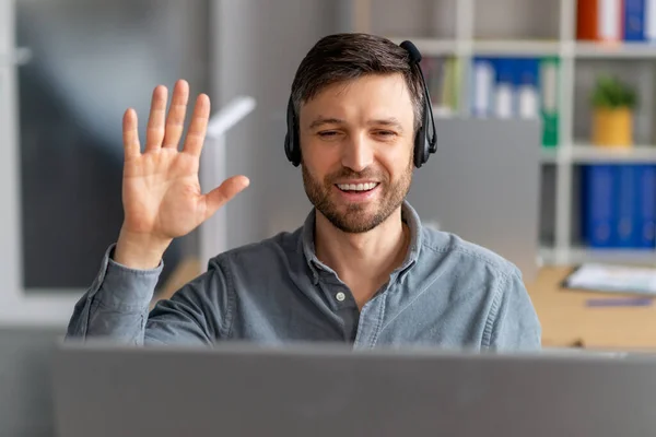 Afstandscommunicatie. gelukkig volwassen man in headset zwaaien hand op computer webcamera terwijl het maken van video call in office — Stockfoto