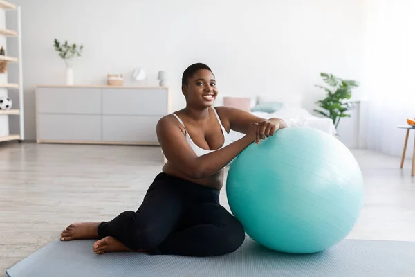 Regordeta joven negra sentada cerca de la pelota de fitness, haciendo ejercicio en la alfombra deportiva en casa —  Fotos de Stock