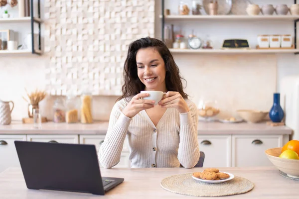 Gelukkig vrouw zitten in de voorkant van laptop aan keukentafel, kijken film op de computer en het drinken van koffie — Stockfoto