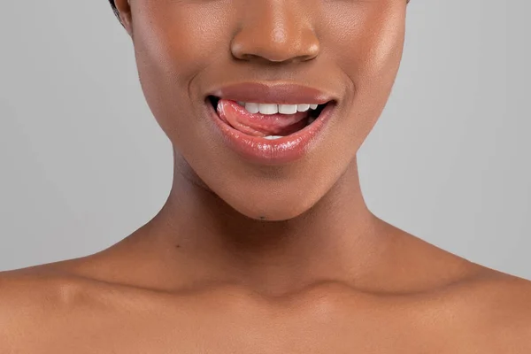 Счастливая африканская женщина тысячелетия с идеальной улыбкой облизывает губы, изолированные на сером фоне — стоковое фото